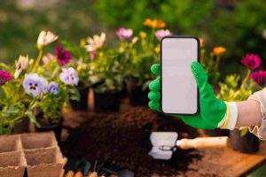A person wearing green gardening gloves holds a smartphone in a garden. Bright flowers and gardening tools are visible, creating a fresh, lively scene