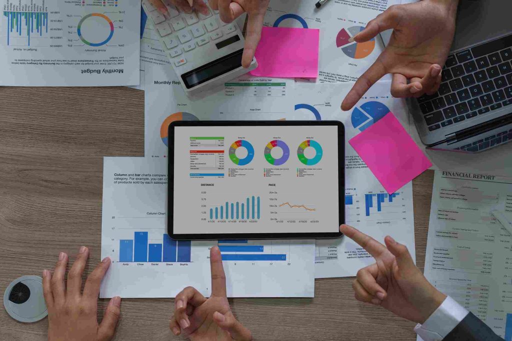 A tablet displaying charts and graphs is surrounded by hands pointing at documents and a laptop on a table, conveying a busy, collaborative meeting.