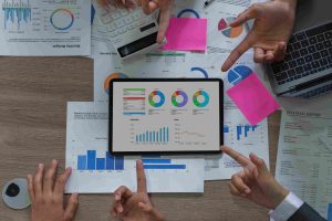 A tablet displaying charts and graphs is surrounded by hands pointing at documents and a laptop on a table, conveying a busy, collaborative meeting.